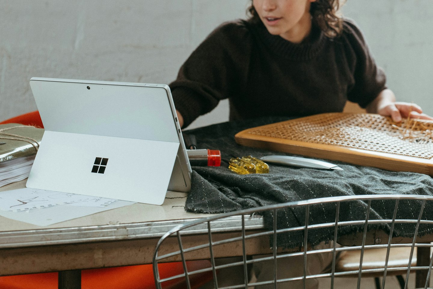 Craftsperson weaving a wooden cane frame next to a laptop.